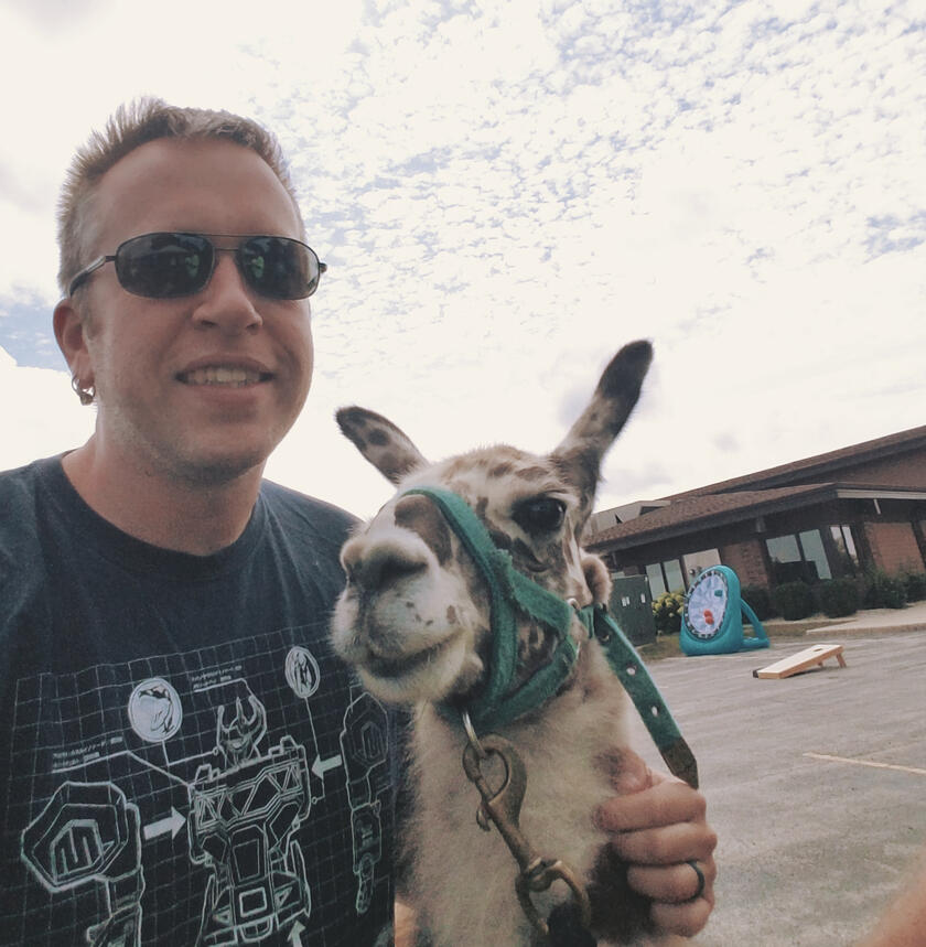 Jeremiah with a llama on a partly cloudy day Jeremiah wearing sunglasses and a graphic t-shirt, standing next to a llama in a parking lot with a partly cloudy sky overhead
